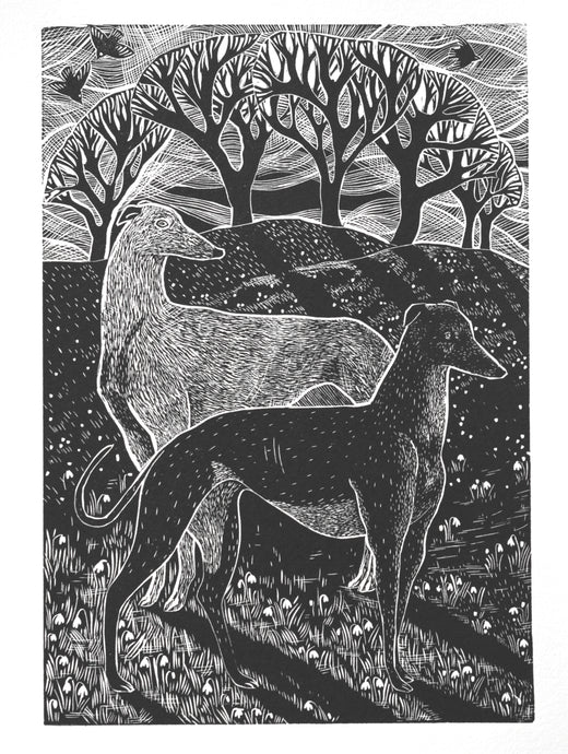 A black and white hand printed linocut art print depicting two greyhounds amidst snowdrops, with tree silhouettes in the background.