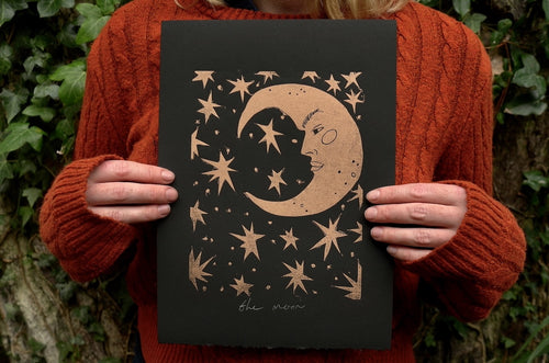 Person holding a black and gold linocut print with a celestial design featuring stars and a moon with a face against a natural background.