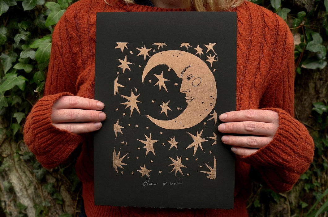 Person holding a black and gold linocut print with a celestial design featuring stars and a moon with a face against a natural background.